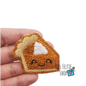 May include: A sparkly orange and brown felt pumpkin pie slice with a white swirl of whipped cream on top. The pie slice has a cute smiling face with black eyes and a small mouth.