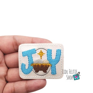 May include: White felt patch with the word "JOY" in blue embroidery. A gold star and a brown manger with a white baby Jesus inside are in the center of the patch.
