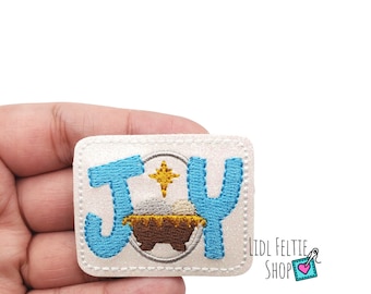 May include: White felt patch with the word "JOY" in blue embroidery. A gold star and a brown manger with a white baby Jesus inside are in the center of the patch.