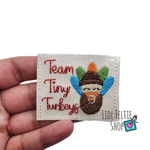 May include: White felt patch with red embroidered text that reads "Team Tiny Turkeys". A cartoon turkey with brown feathers, a blue and green hat, and a blue bib is embroidered on the patch.