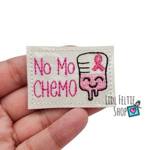 May include: White felt patch with pink embroidery. The patch features a cartoon IV bag with a pink ribbon on top and the text "No Mo Chemo".