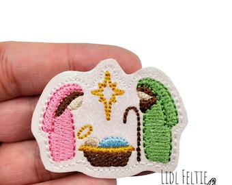 May include: A white felt Christmas ornament with an embroidered nativity scene. The scene includes Mary, Joseph, and baby Jesus in a manger. A gold star is above the manger.