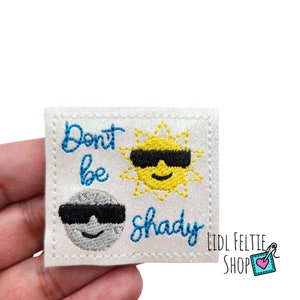 May include: Embroidered patch with the words "Don't be shady" in blue script. The patch features a yellow sun and a gray moon, both wearing black sunglasses. The patch is square with a white background and a glittery border.