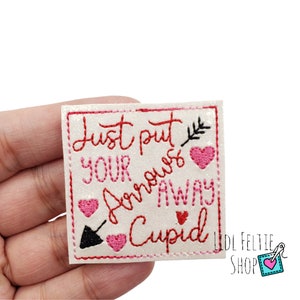 May include: Square, off-white felt patch with red and pink embroidered text and graphics. The text reads "Just put your arrows away Cupid." Features a black arrow, pink hearts, and a small leaf. The patch is held in a hand.