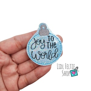 May include: A light blue Christmas ornament-shaped felt applique with the words "Joy to the World" in black script. The ornament has a glittery texture and a gray top. The applique is a festive holiday decoration.