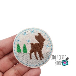 May include: A white felt circle with a brown deer, two green trees, and blue snowflakes embroidered on it. The circle has a blue stitched border and a glittery white background.