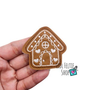 May include: A brown gingerbread house applique with white embroidered details, including hearts and a cross-shaped window. The applique is held in a hand against a white background. The text "LIDL FELTIE SHOP" is visible.