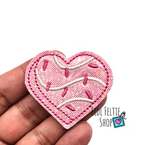 May include: A pink heart-shaped patch with a glittery texture. The patch features white and pink embroidered details resembling sprinkles and icing. The edges are outlined with a darker pink stitching. The text "LIDL FELTIE SHOP" is visible in the lower right corner.