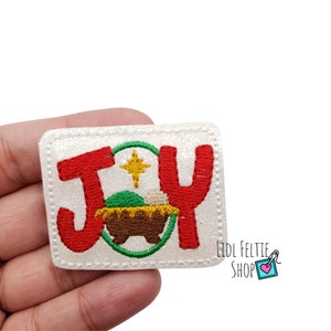 May include: White glitter iron-on patch with red embroidered letters spelling "JOY" and a green and brown embroidered manger scene with a yellow star.