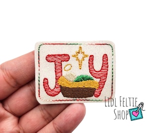 May include: A white felt Christmas ornament with red embroidered letters spelling "JOY" and a gold star above the "O". The "O" is a manger with a baby Jesus inside.