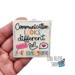 May include: A square, off-white felt patch with the embroidered text "Communication LOOKS different FOR everyone." The text is in black, with the word "LOOKS" in rainbow colors. The patch includes a keyboard, hand sign, and heart symbol.