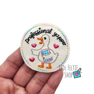 May include: A round, glittery white patch with the words "professional yapper" in black script. The patch features a cartoon duck with a blue and pink design on its chest, surrounded by pink hearts. The patch is held in a hand.