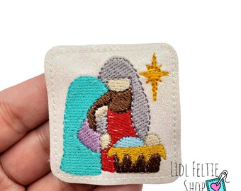 May include: Embroidered patch featuring a Nativity scene with Mary, Joseph, and baby Jesus in a manger. The patch has a glittery white background, with a teal arch, and a gold star. The patch is square with a stitched border.