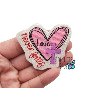 May include: Embroidered patch featuring a pink heart with the word "Love" and a purple cross. The phrase "never fails" is stitched in red. The patch has a white border and is likely for clothing or accessories.