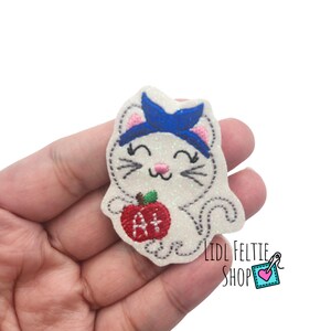May include: A white glittery felt cat applique featuring a blue headband, pink nose, and whiskers. The cat holds a red apple embroidered with the white text A+ on its front. The design is outlined with grey stitching and includes pink inner ears.