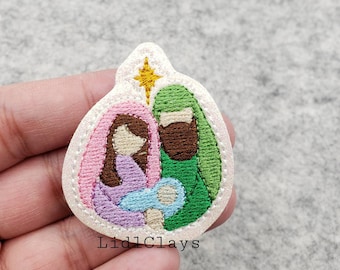 May include: A white felt Christmas ornament with an embroidered image of the Holy Family. The image features Mary, Joseph, and baby Jesus. Mary is wearing a pink and purple robe, Joseph is wearing a green robe, and baby Jesus is wrapped in a blue blanket. A gold star is above the family.