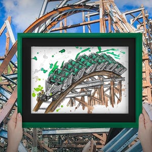 May include: A framed watercolor illustration of a roller coaster with green seats and a wooden track. The artwork is held in front of a weathered roller coaster structure, with a blue sky in the background.