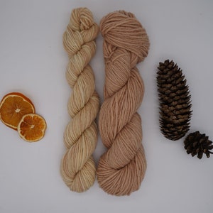 May include: Two skeins of yarn, one in a light beige color and the other in a light peach color. The yarn is arranged in a criss-cross pattern.