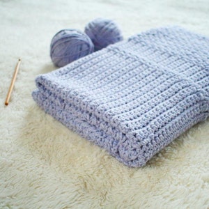 May include: A light purple crocheted blanket folded on a white fluffy surface with two balls of yarn and a crochet hook.