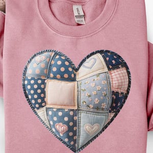 May include: A pink sweatshirt featuring a large, patchwork heart design. The heart is composed of various fabric patterns, including polka dots, floral prints, and gingham, in shades of blue, pink, and white. The sweatshirt has a crew neck and long sleeves.