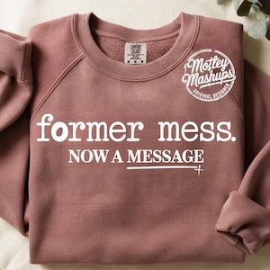 Könnte beinhalten: Mauvefarbenes Sweatshirt mit dem weißen Text "former mess. NOW A MESSAGE". Das Sweatshirt mit Rundhalsausschnitt hat lange Ärmel und ein rundes Logo mit der Aufschrift "Mottey Mashups, Original Designer".