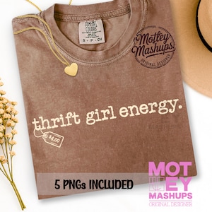 Op de afbeelding: Een bruin T-shirt met de witte tekst "thrift girl energy". Een gouden hartvormige hangerketting en een prijskaartje zijn zichtbaar. Het shirt is van Comfort Colors en heeft een klein maatlabel. Een hoed en bloemen staan op de achtergrond.