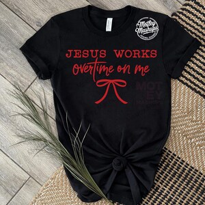 May include: Black t-shirt featuring the red text "JESUS WORKS overtime on me" and a red bow graphic. The shirt is knotted at the hem. The brand logo "Motley Mashups" is in the upper right corner.