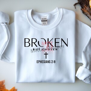 May include: White sweatshirt with the text "BROKEN but CHOSEN" in black and pink, a cross, and "Ephesians 2:8". The long-sleeved top is folded on a light surface.