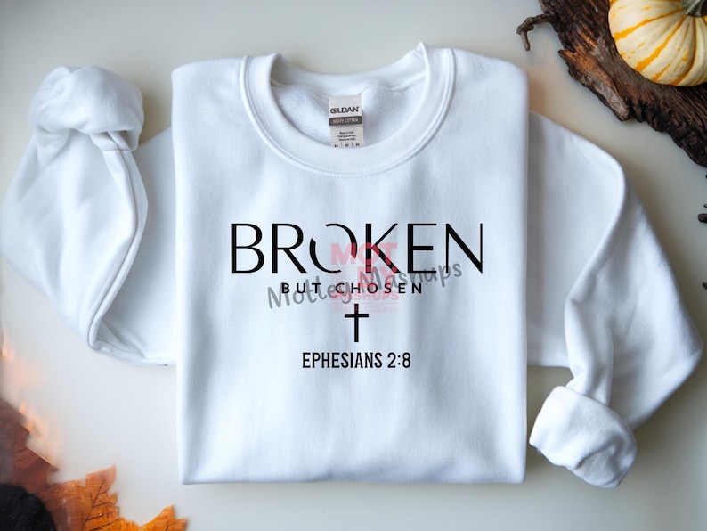 May include: White sweatshirt with the text "BROKEN but CHOSEN" in black and pink, a cross, and "Ephesians 2:8". The long-sleeved top is folded on a light surface.