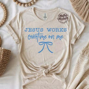 May include: A cream-coloured t-shirt featuring the blue text "JESUS WORKS overtime on me" and a blue bow illustration. The shirt is knotted at the waist. The brand logo "Motley Mashups" is visible on the upper right.