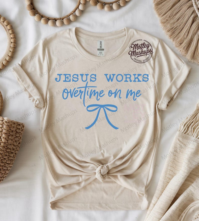 May include: A cream-coloured t-shirt featuring the blue text "JESUS WORKS overtime on me" and a blue bow illustration. The shirt is knotted at the waist. The brand logo "Motley Mashups" is visible on the upper right.