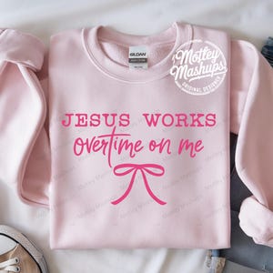 May include: A pale pink sweatshirt featuring the phrase "JESUS WORKS overtime on me" in pink lettering, with a pink bow graphic. The sweatshirt has a crew neck and long sleeves. The brand name "Mottey Mashups" is in a circle on the top right.