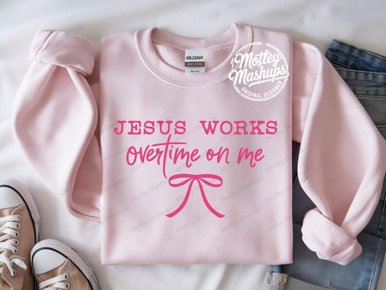 May include: A pale pink sweatshirt featuring the phrase "JESUS WORKS overtime on me" in pink lettering, with a pink bow graphic. The sweatshirt has a crew neck and long sleeves. The brand name "Mottey Mashups" is in a circle on the top right.
