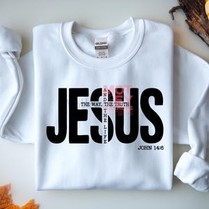 May include: White sweatshirt with the word "JESUS" in large black letters. The words "NOT" and "THE WAY, THE TRUTH, THE LIFE" are in a smaller, pink font. The text "JOHN 14:6" is at the bottom in black.
