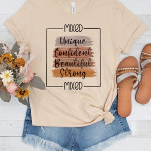 Könnte beinhalten: Ein beiges T-Shirt mit einem grafischen Design, das "Mixed" über einem Rechteck mit den Worten "Unique", "Confident", "Beautiful" und "Strong" in schwarzer Schrift auf einem Hintergrund aus braunen, orangefarbenen und grauen Pinselstrichen sagt. Die Worte "Mixed" sind unter dem Rechteck geschrieben.
