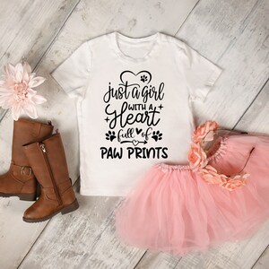 May include: White t-shirt with a black graphic that says "just a girl with a heart full of paw prints". The shirt is on a white wooden surface with a pink tutu and brown boots.