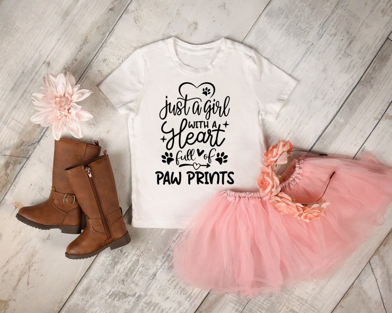 May include: White t-shirt with a black graphic that says "just a girl with a heart full of paw prints". The shirt is on a white wooden surface with a pink tutu and brown boots.