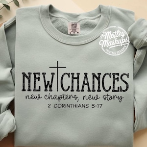 May include: A light green sweatshirt with the words "NEW CHANCES" in a bold, black font, with a cross above the text. Below, it reads "new chapters, new story" and "2 CORINTHIANS 5:17". The sweatshirt has a crew neck and long sleeves.