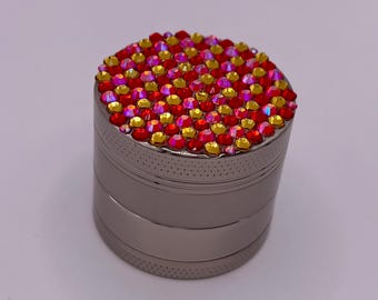 Op de afbeelding: Een zilveren metalen grinder met een bovenkant bedekt met rode en gouden strass-steentjes. De grinder heeft een cilindrische vorm met meerdere secties. De bovenkant is volledig ingelegd met de kleurrijke strass-steentjes, wat een sprankelend effect creëert.