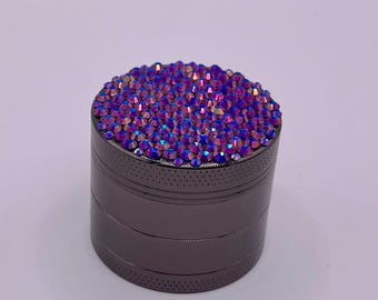Op de afbeelding: Een donkergrijze, cilindrische grinder met een bovenoppervlak volledig ingelegd met iriserende paarse en blauwe kristallen. De grinder heeft een geribbeld ontwerp en kleine ventilatiegaten.