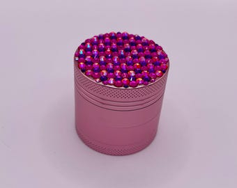 Op de afbeelding: Een roze metalen grinder met een bovenkant bedekt met levendige roze en paarse strass-steentjes. De grinder heeft een cilindrische vorm met een textuur en kleine gaatjes in het midden.