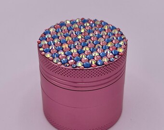 Op de afbeelding: Een roze, cilindrische grinder met een bovenkant bedekt met kleurrijke strass-steentjes. De strass-steentjes zijn een mix van blauwe, roze en iriserende kleuren. De grinder heeft een gestructureerd, geperforeerd ontwerp rond de boven- en onderkant.