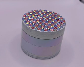 Op de afbeelding: Een cilindrische, iriserende grinder met een deksel volledig ingelegd met kleine, kleurrijke strass-steentjes. De body van de grinder heeft een parelmoer afwerking, die tinten roze, blauw en wit reflecteert. De strass-steentjes zijn een mix van roze, blauw en helder.