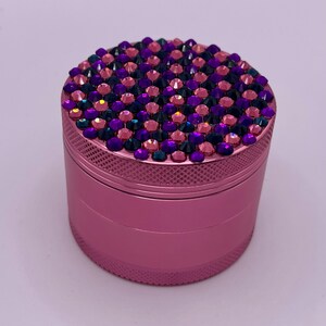 May include: A pink, cylindrical grinder with a textured exterior. The top is fully encrusted with a mosaic of pink, purple, and dark blue rhinestones. The grinder is designed for herbs and spices.
