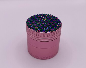 Op de afbeelding: Een roze, cilindrische grinder met een bovenkant bedekt met iriserende, juweelkleurige strass-steentjes. De grinder heeft een gestructureerd, geperforeerd ontwerp rond de boven- en onderranden.