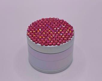 Op de afbeelding: Een cilindrische, zilverkleurige grinder met een bovenkant volledig ingelegd met kleine, roze strass-steentjes. De behuizing van de grinder heeft een lichtroze, iriserende afwerking. De grinder is ontworpen voor kruiden en specerijen.