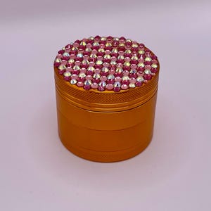 May include: A gold-colored, cylindrical grinder with a textured surface. The top is fully encrusted with pink and iridescent rhinestones. The grinder is designed for herbs and spices.