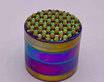 Op de afbeelding: Een cilindrische grinder met een regenboogkleurige iriserende afwerking. De bovenkant is bedekt met kleine, groene en bruine strass-steentjes. De grinder heeft een metallic glans en een veelkleurige, horizontale streep.