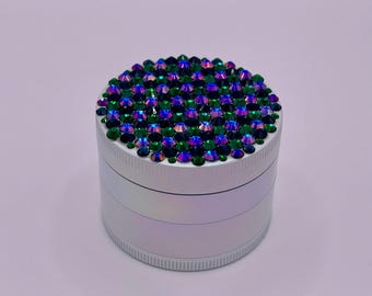 Op de afbeelding: Een zilveren grinder met een bovenkant bedekt met iriserende groene en blauwe strass-steentjes. De grinder heeft een cilindrische vorm met een geribbelde bodem en een glad, reflecterend oppervlak.
