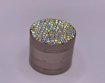 Op de afbeelding: Een zilverkleurige grinder met een bovenoppervlak volledig ingelegd met iriserende strass-steentjes. De grinder heeft een cilindrische vorm met horizontale groeven en een geperforeerde band aan de bovenkant. De strass-steentjes reflecteren een regenboog aan kleuren.